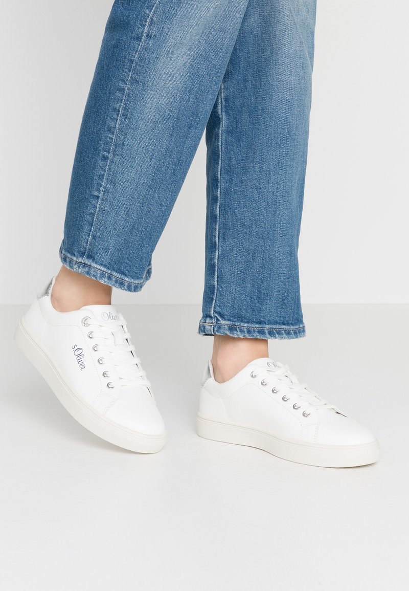 Weiße Sneaker mit Obermaterial aus Leder, silbernen Akzenten und sechs Ösen. Getragen mit hochgerollten blauen Jeans, die ein lässiges Design zur Schau stellen.