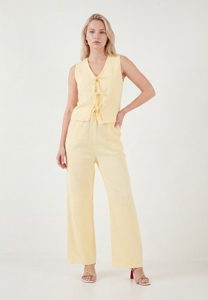 Naine seisab, kandes varrukateta kahvatukollast topi ees sõlmedega ning sobivaid laiade säärtega pükse, jalas avatud varbaga kontsakingad, valgel taustal.