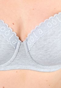 Harmaa rintaliivi, jossa on aaltoileva pitsireuna ja painettu teksti. Pehmeä kangas ja sileä pinta, keskellä sauma.