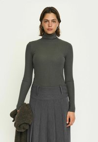 Dunkelgrünes, geripptes Rollkragenoberteil mit langen Ärmeln, kombiniert mit einem grauen, plissierten Rock. Ein dunkler Pullover ist über den linken Arm gelegt.