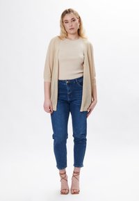 Cárdigan abierto de punto beige sobre una blusa beige claro, combinado con jeans de tiro alto azul y tacones desnudos con tiras, de pie contra un fondo liso.