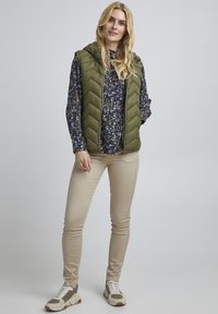 Grøn quiltet vest med hætte over en blå blomster langærmet skjorte, beige slim bukser og hvide sneakers med nøddeagtige detaljer.