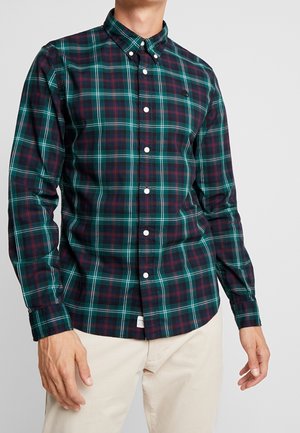 Mann trägt ein dunkelgrünes, marineblaues und rotes kariertes Hemd mit weißen Knöpfen und beigen Hosen, steht vor einem einfarbigen Hintergrund.