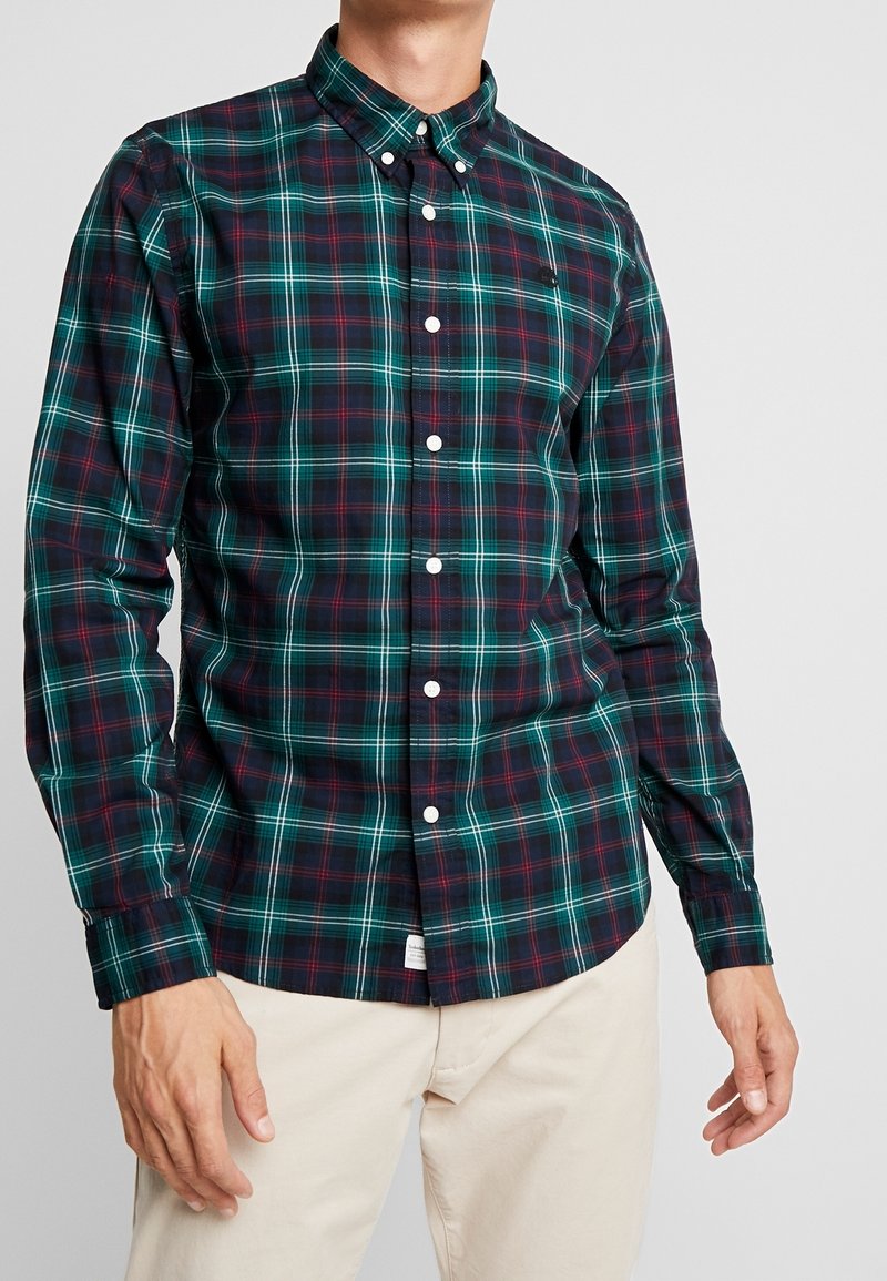 Homme portant une chemise à carreaux bleu marine, vert foncé et rouge avec des boutons blancs, ainsi qu'un pantalon beige, debout devant un fond uni.