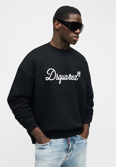 Άνδρας που φοράει μαύρο φαρδιά φούτερ με το λογότυπο "Dsquared²", γυαλιά ηλίου τύπου wraparound με ανοιχτόχρωμους φακούς και ανοιχτόχρωμα μπλε τζιν με τα χέρια στις τσέπες.