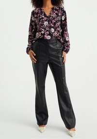 Žena v černých kožených kalhotách, květinové blůze s dlouhým rukávem a bílých špičatých podpatcích stojící na neutrálním pozadí.