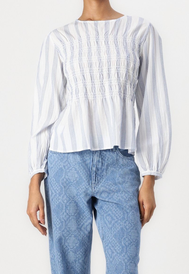 Haut blanc avec des rayures verticales bleues, corsage froncé et manches bouffantes, associé à un jean bleu clair à motif serpent.