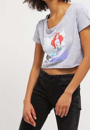 Vrouw met een grijs cropped t-shirt met een kleurrijke zeemeerminprint en een zwarte high-waisted jeans tegen een effen achtergrond.