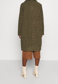 Olivengrønt struktureret frakke med stor krave, midtlængde design, båret over brune ruskinds shorts og cremede ankelstøvler med kraftige hæle.