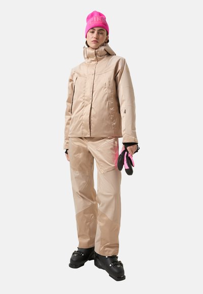 Beige skijas en broek met een gladde textuur. Roze gebreide muts en zwarte handschoenen. De laarzen zijn zwart met gespen.