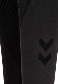 Zbliżenie czarnej teksturowanej tkaniny sportowej z dwoma ciemnymi paskami w kształcie szewrona na rękawie lub nogawce spodni.