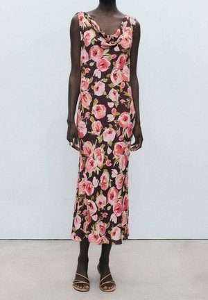 Mujer con un vestido midi marrón sin mangas con estampado floral rosa y sandalias beige con tiras, de pie frente a un fondo liso.