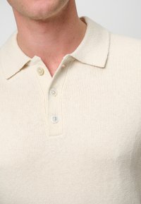 Beige gestricktes Polohemd mit Kragen, drei weißen Knöpfen und einer strukturierten Oberfläche, die ein dezentes horizontales Rippmuster aufweist.