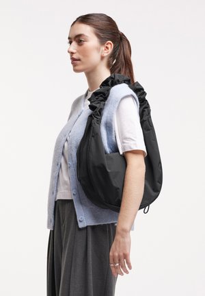 Junge Frau mit Pferdeschwanz, die einen hellblauen Cardigan, ein weißes T-Shirt und einen grauen Rock trägt und eine große schwarze geraffte Schultertasche auf der rechten Schulter trägt.