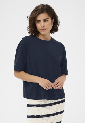 Femme aux cheveux bruns courts et ondulés portant un haut ample à manches courtes bleu foncé et une jupe blanche avec des rayures horizontales bleu marine.