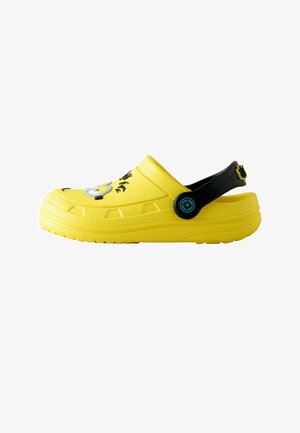 Zoccolo slip-on giallo con cinturino posteriore nero regolabile, con design del volto di un Minion sulla parte superiore e suola testurizzata per la presa.