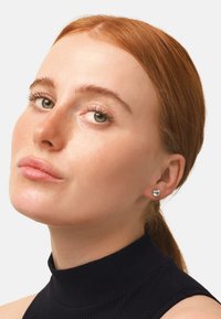 Silberne Steckerohrringe mit einem klaren, runden Stein, der in einem schlichten, polierten Metall gefasst ist. Tragen von einem Model mit roten Haaren.
