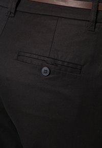 Gros plan sur un pantalon noir avec une poche arrière boutonnée et une ceinture marron visible à la taille.