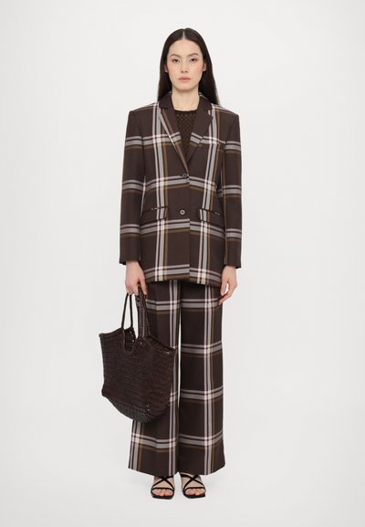 Completo a quadri marrone con pantaloni a gamba larga e una giacca su misura. Presenta strisce beige, bianche e grigie. Il modello tiene una borsa a tracolla in tessuto scuro.