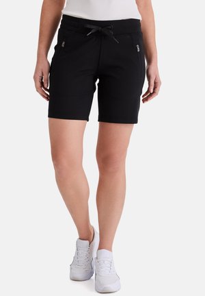 Frau trägt schwarze Shorts mit Kordelzug und seitlichen Reißverschlusstaschen sowie weiße Turnschuhe, steht vor weißem Hintergrund.