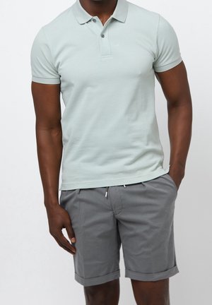 Man wearing light green short-sleeve polo shirt and gray rolled-hem shorts with hands in pockets against plain background.