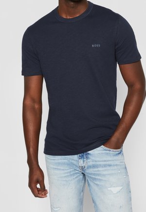 Mężczyzna ubrany w granatowy ciemny t-shirt z krótkim rękawem z napisem „BOSS” na piersi oraz jasnoniebieskie, przetarte dżinsy, z jedną ręką w kieszeni.
