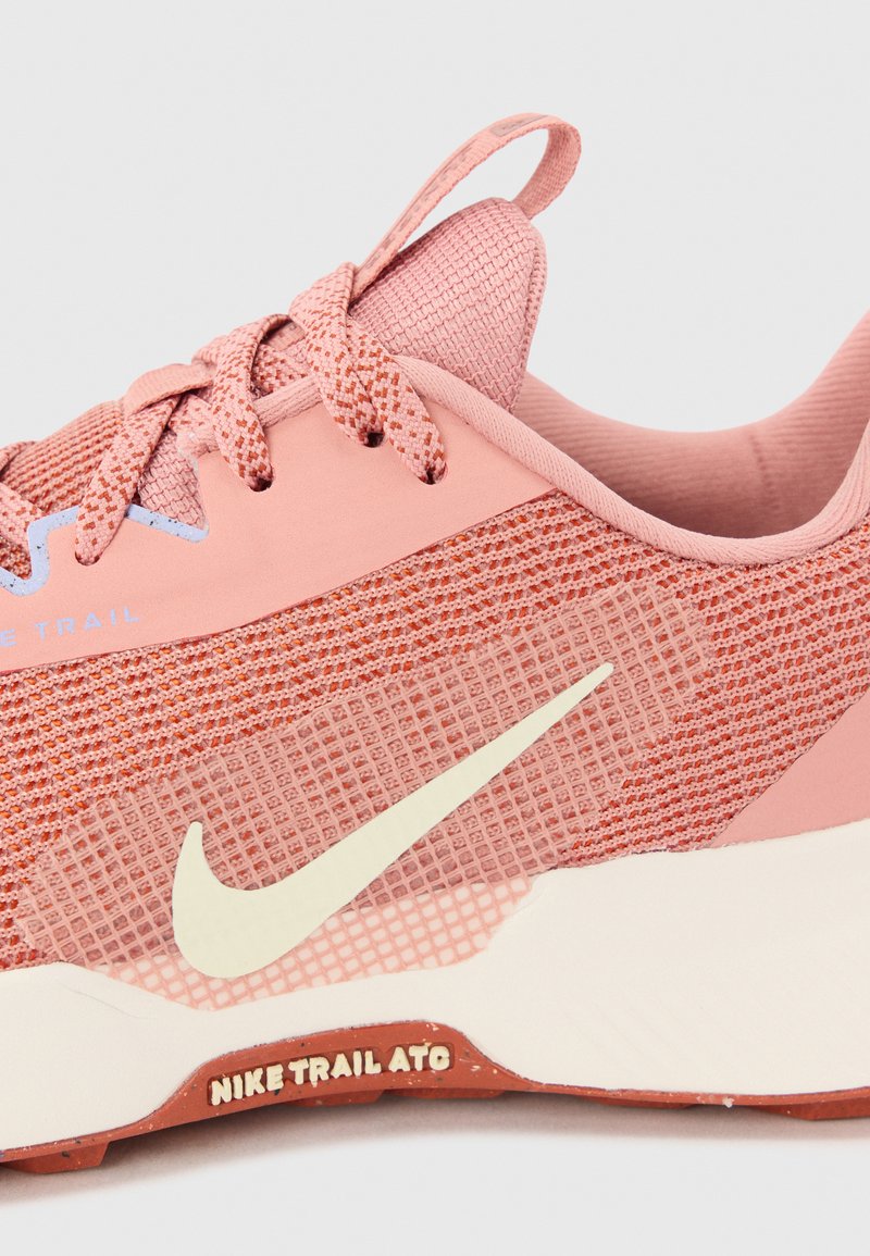Jasnoróżowe buty do biegania w terenie marki Nike, z teksturowaną siateczką, kontrastującą jasną podeszwą oraz wyraźnym logo Swoosh w kolorze kremowym. Posiadają sznurówki oraz pętelkę do zakładania.