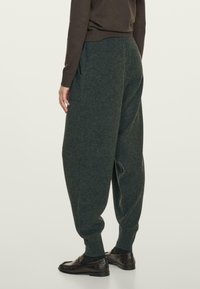 Persoon in donkergrijze losse tapered broek, donkere bruine schoenen en een donkerbruine blouse met lange mouwen, staand tegen een effen lichte achtergrond.
