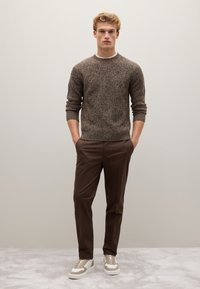 Maglione marrone lavorato a maglia con un motivo testurizzato, abbinato a pantaloni marroni su misura e scarpe da ginnastica crema con dettagli marroni.