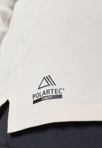 Tkanina boje kreme s glatkom teksturom na kojoj se nalazi POLARTEC® Delta™ logo u crnoj boji. Rub je blago zakrivljen s vidljivim šavovima.