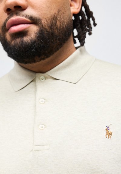 Άτομο με γενειάδα που φοράει μπεζ πόλο μπλούζα με κουμπιά και μικρό κεντητό λογότυπο παίκτη πόλο πάνω σε άλογο.