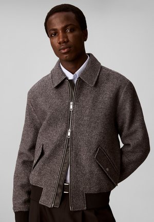 Joven con una chaqueta gris de lana con cremallera sobre una camisa blanca con cuello, de pie con las manos detrás de la espalda frente a un fondo liso.