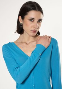 Blauer Cardigan mit V-Ausschnitt, langen Ärmeln, gerippten Bündchen und einer Knopfleiste mit dekorativem Detail am Ärmel. Weicher Stoff.