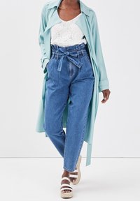 Trench-coat bleu clair sur un haut en dentelle blanche, pantalon en denim bleu taille haute avec ceinture à nouer, et sandales compensées blanches à brides.