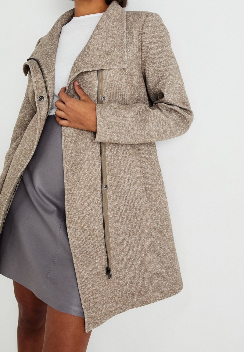 Femme portant un manteau en laine beige sur un haut blanc et une jupe grise, tenant le revers du manteau près de la poitrine sur un fond uni.