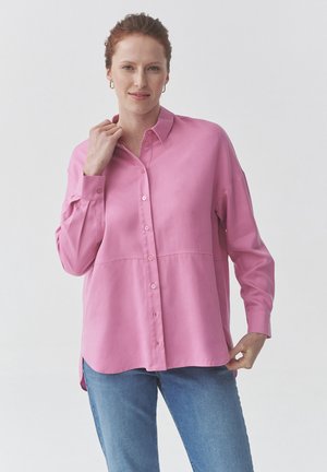 Pink button-up shirt with long sleeves, featuring a slightly relaxed fit and a smooth texture, paired with light blue jeans.