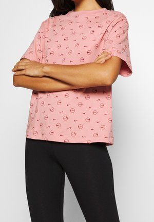 Person wearing pink Nike T-shirt with red logo pattern and black pants, arms crossed across chest, against white background.