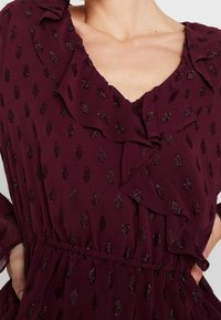 Abito color bordeaux con scollatura volant e sottili motivi a foglia. Tessuto trasparente con accenti luccicanti. Vita elasticizzata; maniche lunghe.
