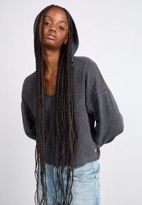 Sweatshirt à capuche gris avec un tissu texturé, coupe ample et épaules tombantes. Assorti à un jean bleu clair. Longs cheveux tressés partiellement visibles.
