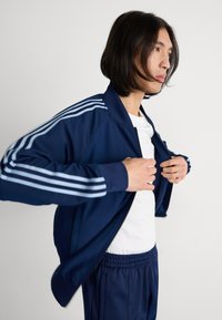 Sötétkék sportdzseki világoskék csíkokkal az ujjakon, cipzáras záródás, bordás mandzsetták, fehér inggel és azonos színű sötétkék nadrággal párosítva.