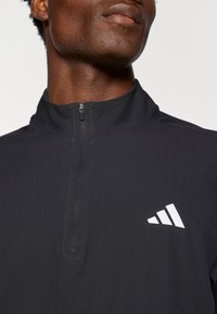 Čierny športový polovičný zips s rebrovanou textúrou. Obsahuje krátky zips a biely logový akcent na ľavej hrudi.