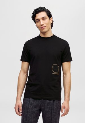 Un mannequin masculin porte un tee-shirt noir avec le logo doré Karl Lagerfeld et un pantalon foncé sur un fond clair uni.
