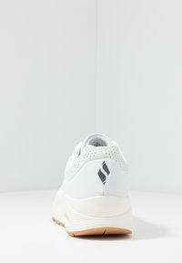 Baskets blanches avec un dessus lisse en synthétique, des détails perforés au niveau du talon et une semelle en caoutchouc avec une semelle extérieure beige.