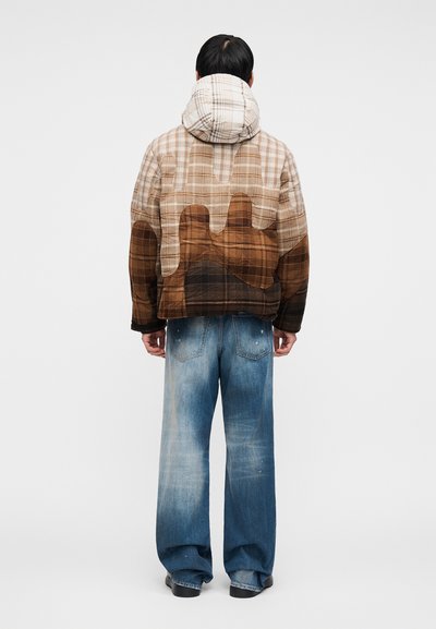 Καπιτονέ μπεζ και καφέ καρό μπουφάν με κουκούλα, με σχέδιο λιώματος στο πίσω μέρος. Συνδυασμένο με φαρδιά τζιν παντελόνια σε ξεθωριασμένο μπλε.