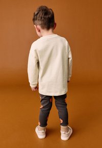 Krafti pitkähihaine vaaleanebene paita, mustat legginsit kõrvitsakuvandiga, beežid sokid, heledad jalanõud; nähtud tagant pruuni tausta ees.