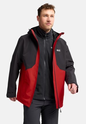 Man die een zwart-rode Jack Wolfskin outdoorjas draagt over een zwarte fleece met rits, staand tegen een effen lichte achtergrond.