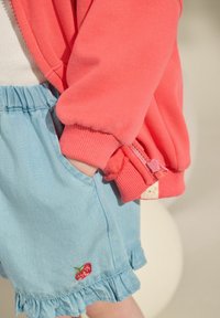 Koraliowy sweter z zamkiem błyskawicznym i ściągaczami na rękawach, w połączeniu z jasnoniebieskimi szortami dżinsowymi z falbaniastym dołem i haftem czerwonej truskawki.