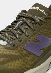 Zapatilla deportiva verde y marrón con parte superior de malla, suela gruesa, logotipo morado, cordones texturizados y acolchado en el tobillo.