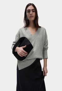 Lichtgrijze, oversized trui met een V-hals en geribbelde details, gecombineerd met een zwarte gehaakte clutch. De outfit bevat een zwarte rok.