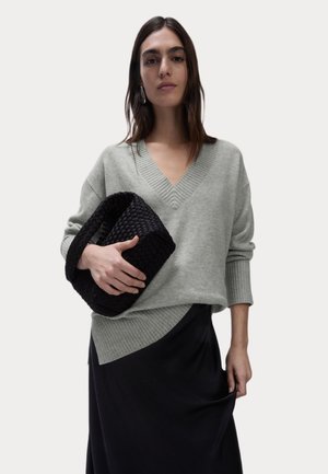Suéter gris claro, de gran tamaño, con escote en V y detalles acanalados, combinado con un bolso de mano negro tejido. El atuendo incluye una falda negra.
